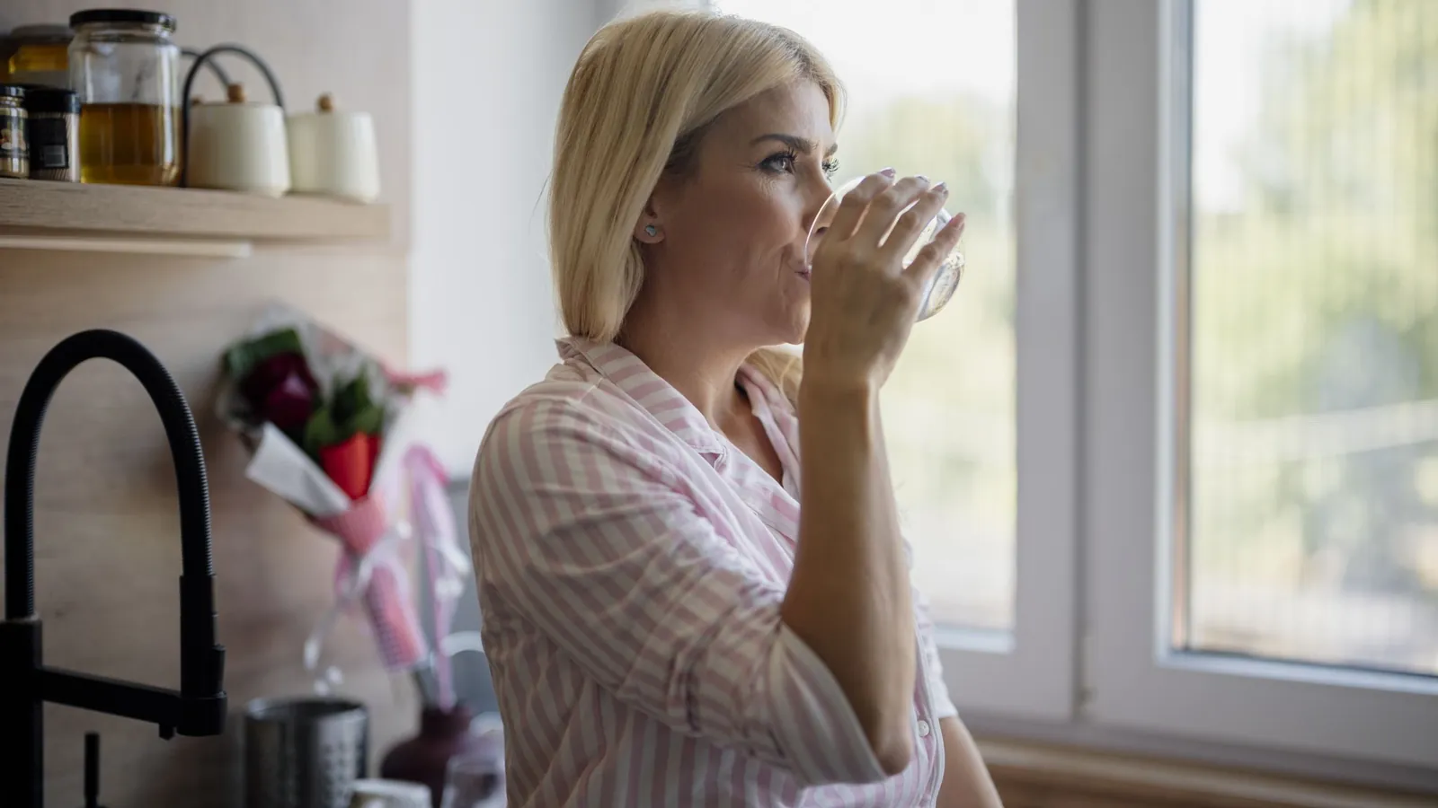 Woman drinking a glass of water by a kitchen window