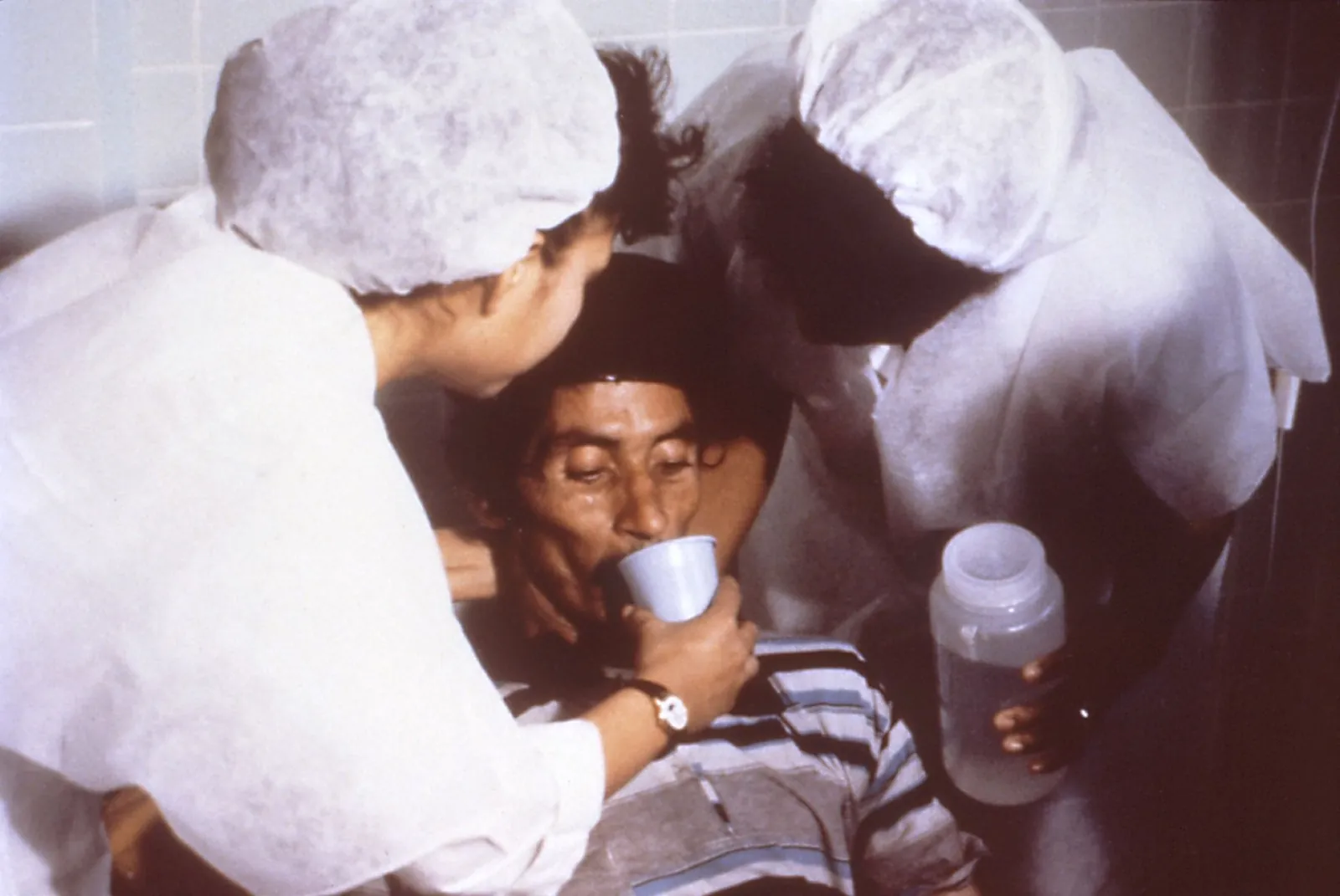 Two nurses helping a patient drink oral rehydration solution from a cup during cholera treatment